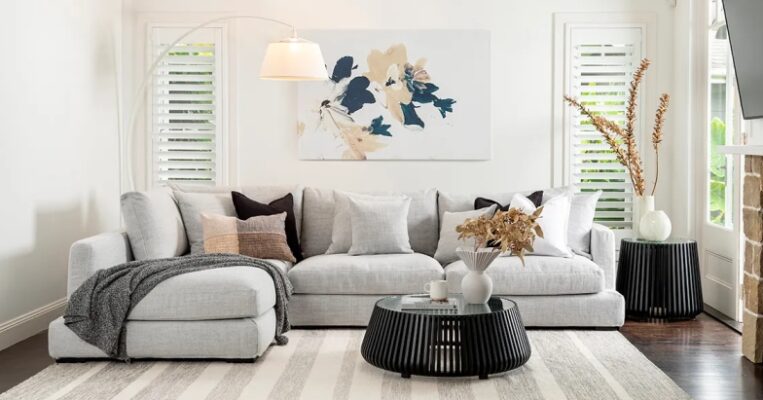 Bright living room with a large light-grey sectional sofa, striped rug, slatted black round coffee table, and abstract floral wall art.